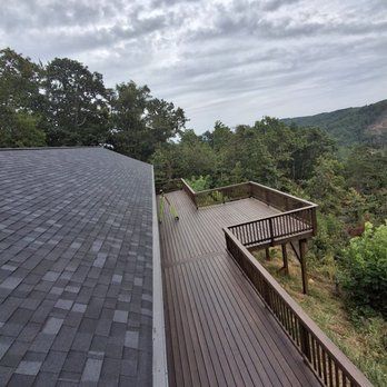 View of a wooden deck extending from a house roof, overlooking a lush green hillside under a cloudy sky.