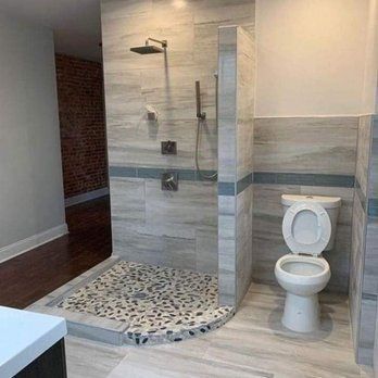A modern bathroom with a tiled shower and toilet. The shower has a pebble floor and a rain shower head. The toilet seat is up.