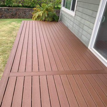 Wooden deck extending from a house with sliding glass doors. The deck is brown with a rectangular section in the middle. Green grass and a wall are visible in the background.