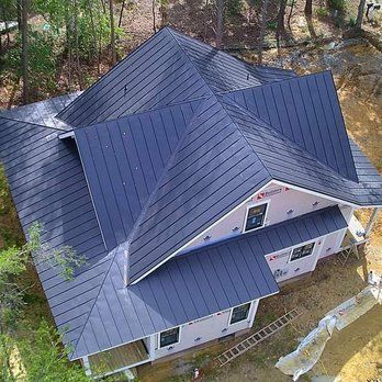 Aerial view of a house with a dark gray metal roof and light pink exterior walls, surrounded by trees.