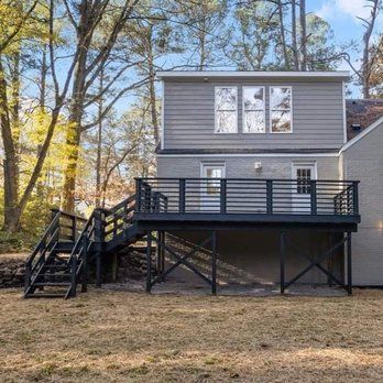 A two-story house with a black deck, stairs, and gray siding. The deck overlooks a grassy yard, with trees in the background.