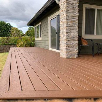 A brown composite deck extends from a green house with a stone pillar. A black chair is on the deck.