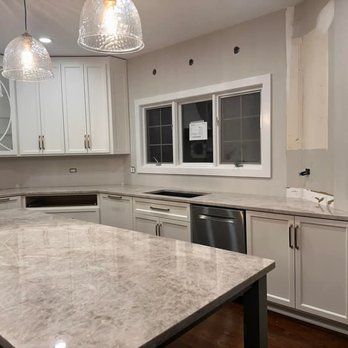 A kitchen with a large island and white cabinetry. A stainless steel dishwasher and a window are visible. The countertops are gray.