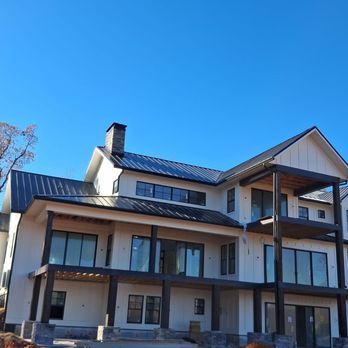 Modern two-story white house with black trim, roof, and chimney, against a clear blue sky. Large windows and a covered patio area.