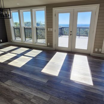 Spacious room with large windows and glass doors opening to a deck overlooking a scenic view. Sunlight streams across the gray wood-look flooring.