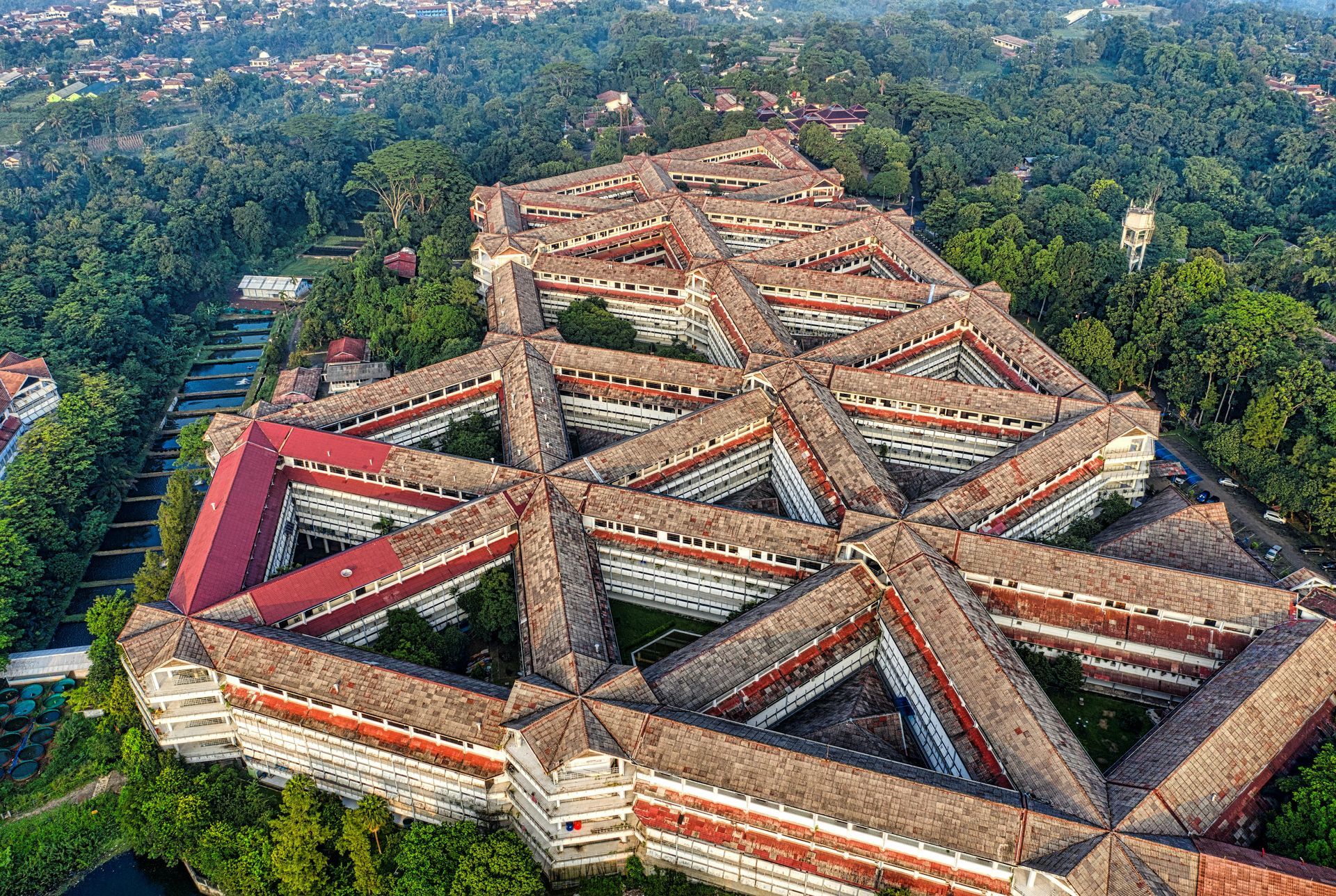 Aerial view of a large, decaying multi-building complex with a complex geometric design, surrounded by lush green trees. Red roofs and white walls.