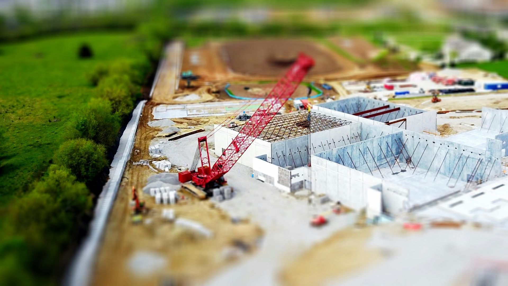 Construction site with a red crane; unfinished white buildings, dirt, and a green field.
