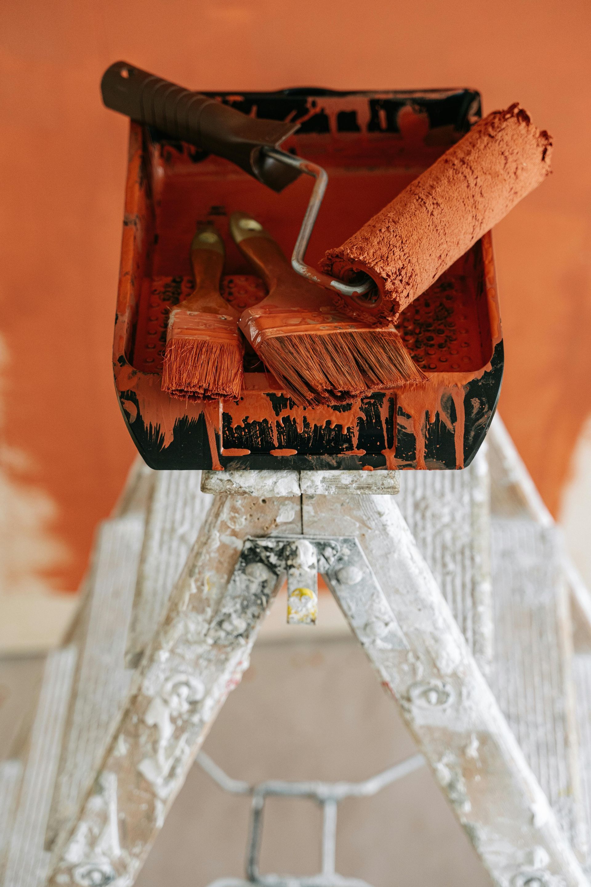 Paint tray with roller, paintbrushes, and paint, atop a step ladder, all covered in orange paint.