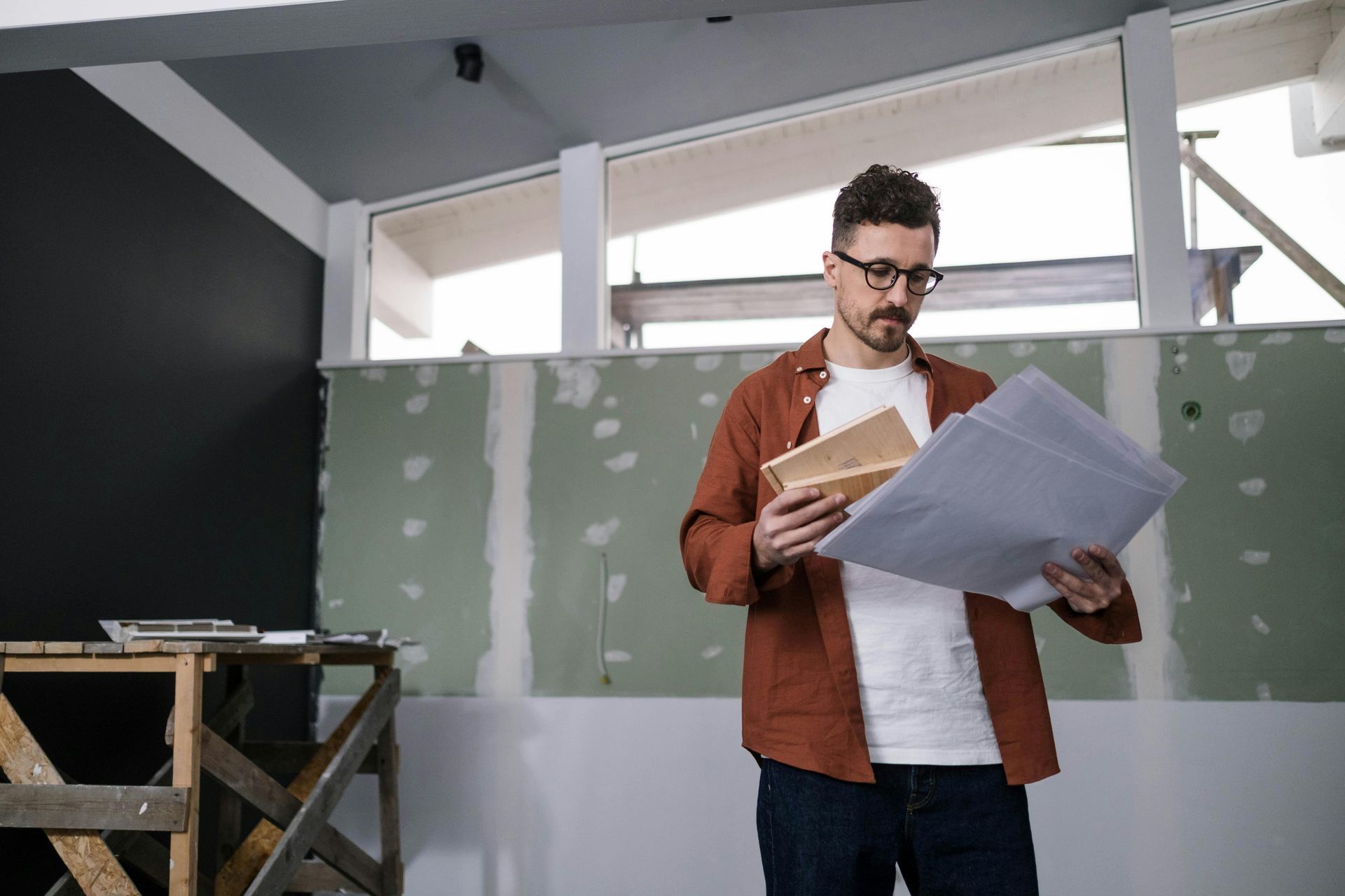 Man in glasses and an orange shirt looks at blueprints and a small box in a room under renovation.