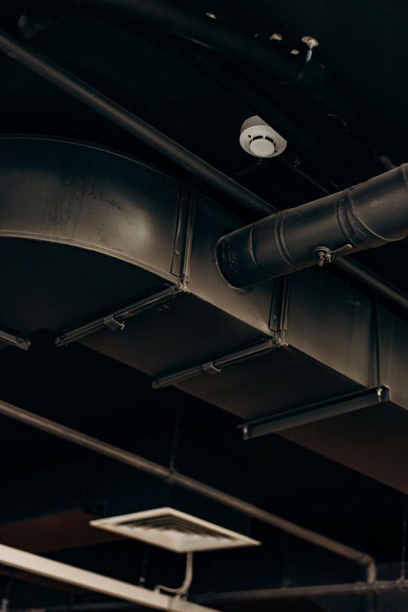Black ceiling with industrial-style ductwork, pipes, and a security camera. A square vent is visible below.