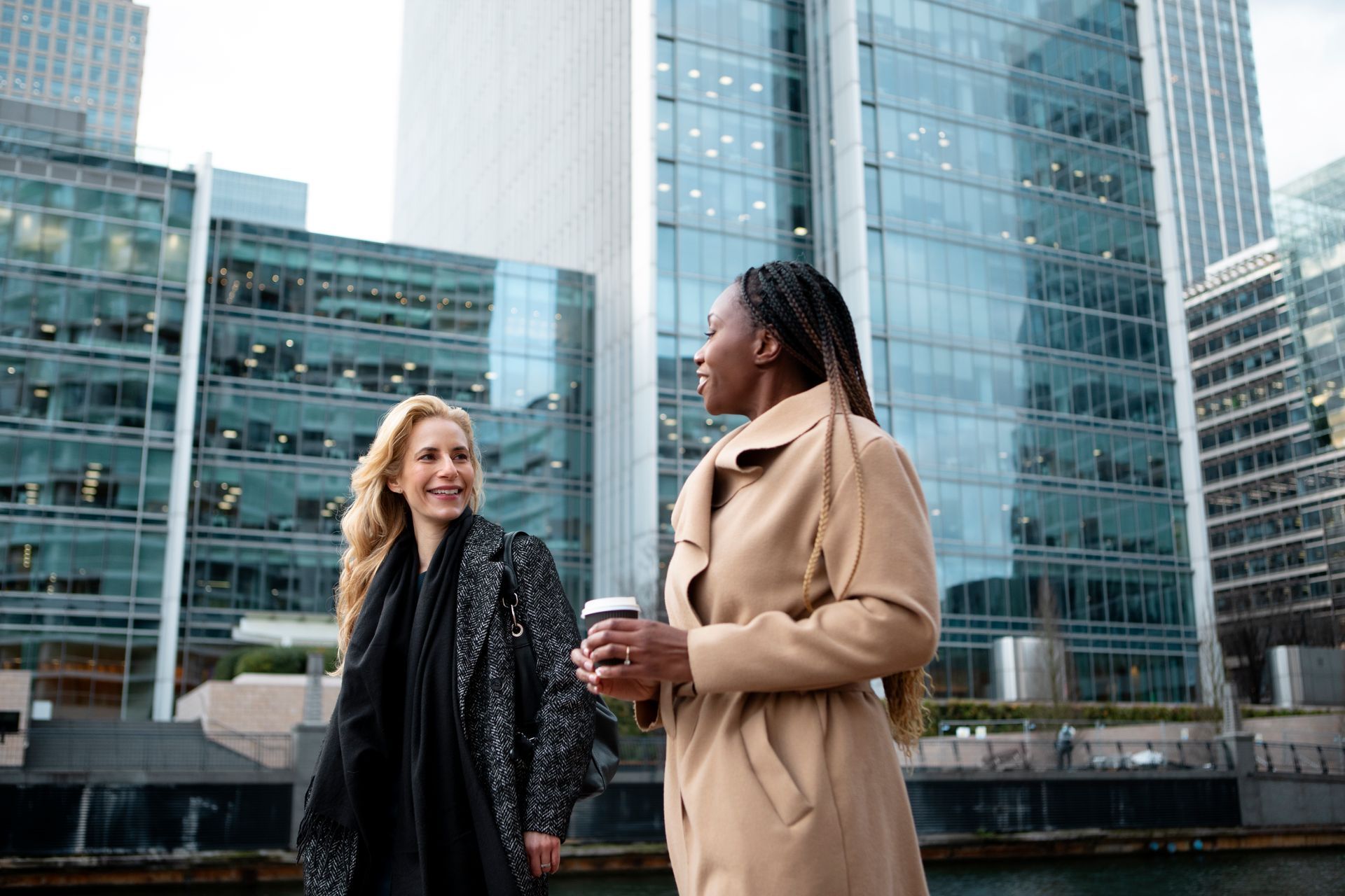 Two women are standing next to each other in front of tall buildings in a city.