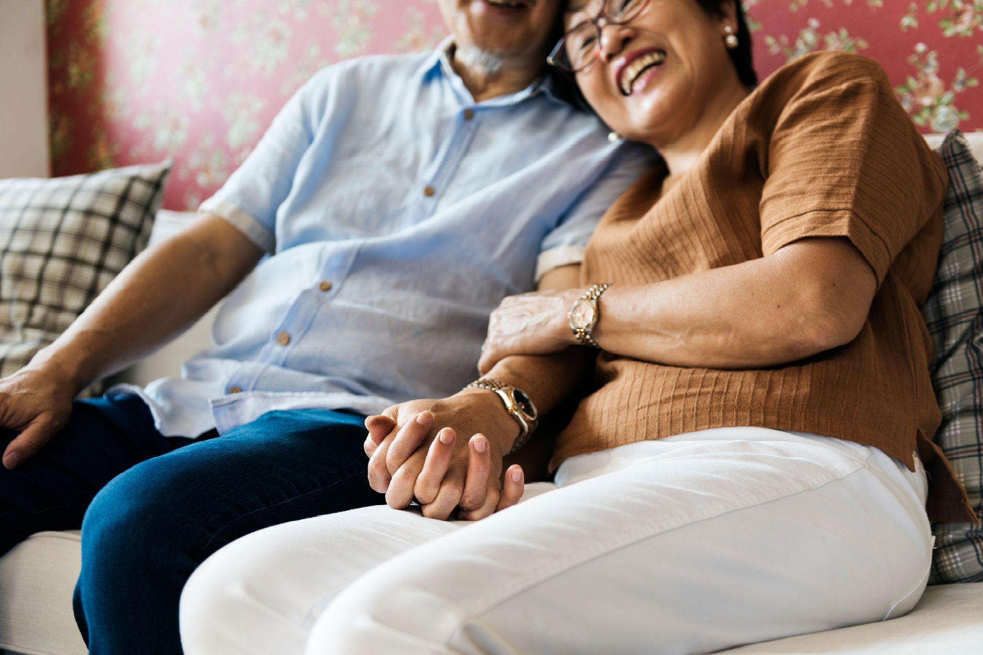 An elderly couple is sitting on a couch holding hands and smiling.