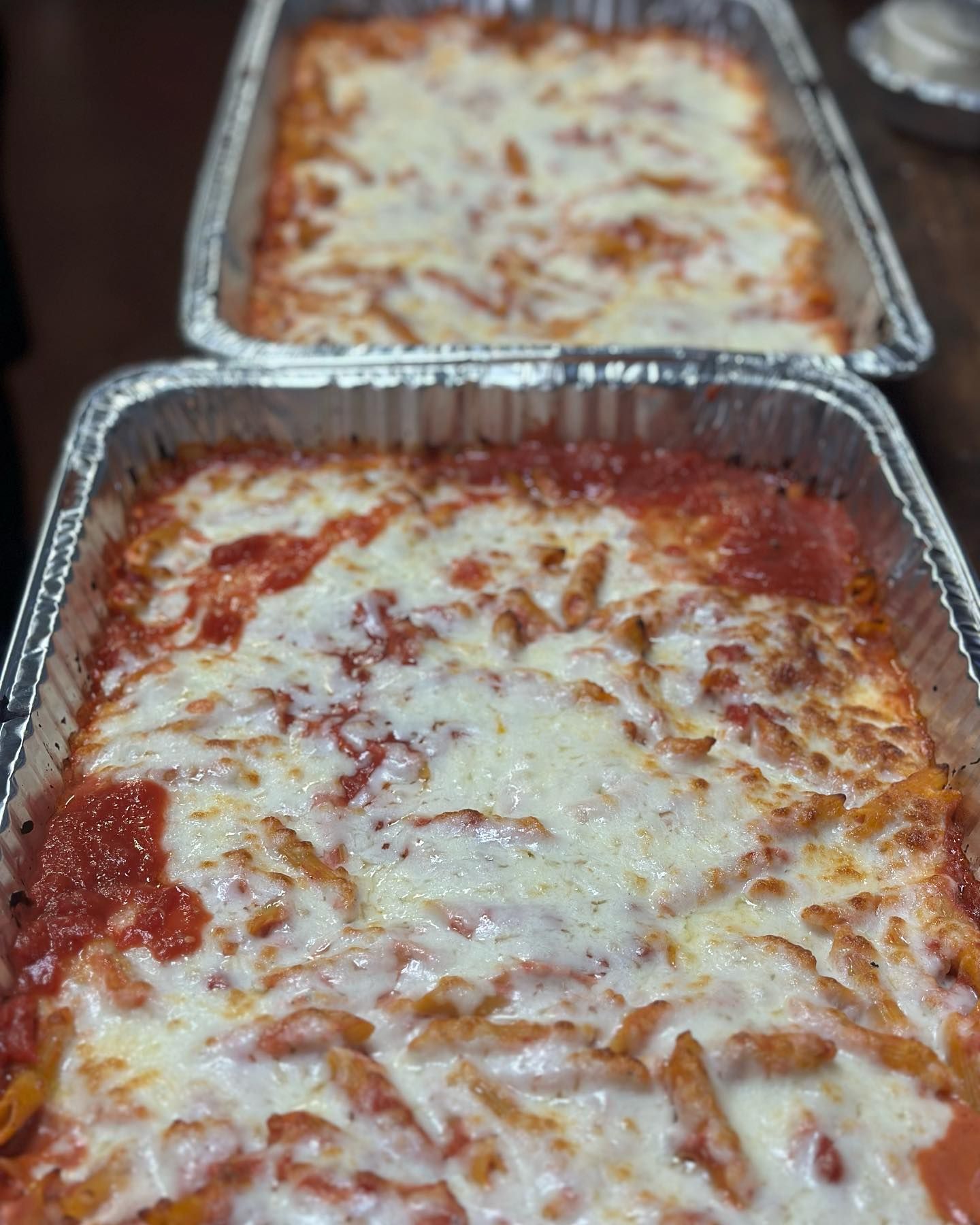 Two rectangular foil trays filled with baked ziti, topped with red sauce and melted cheese.