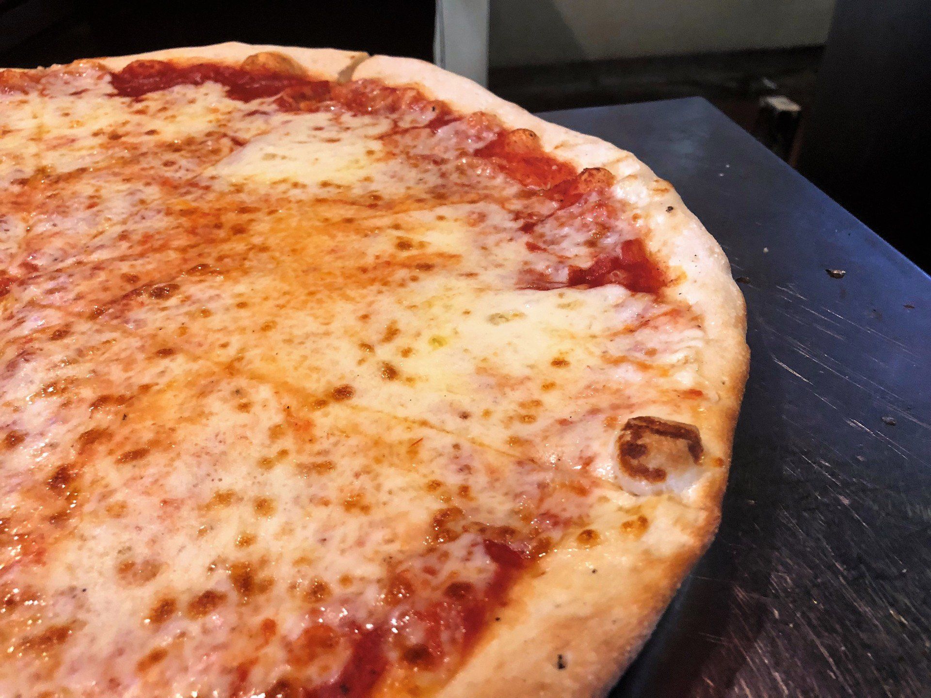 Cheese pizza on a metal surface, close-up. Crust is golden brown with melted white cheese and red sauce.