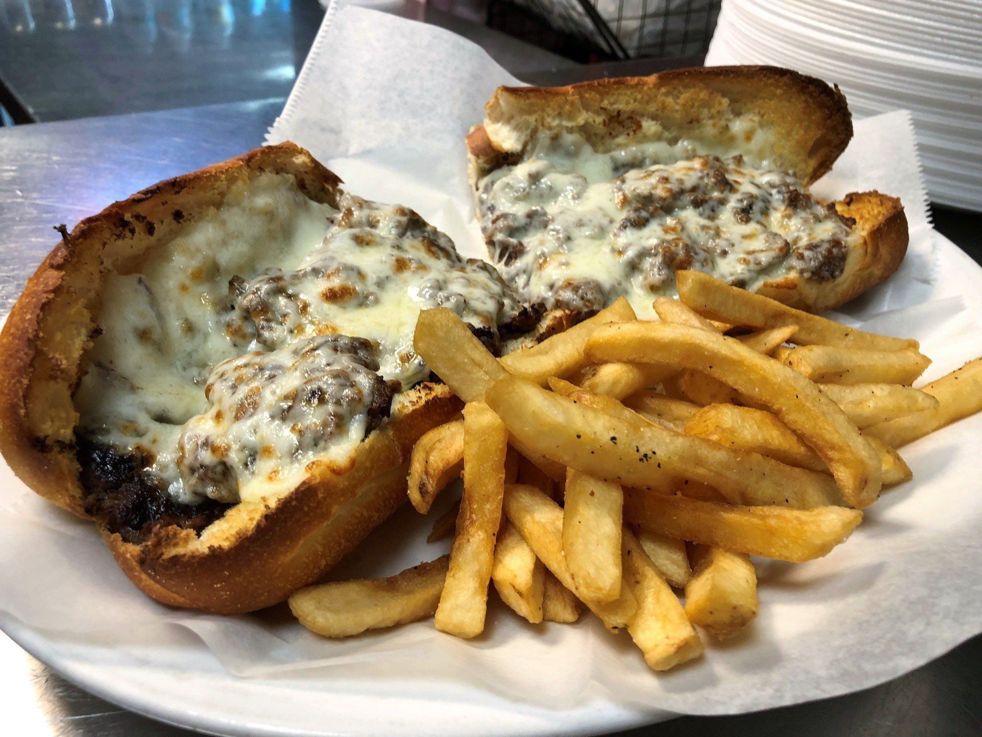 Cheesesteak sandwich with fries on a white plate. The sandwich is cut in half and topped with melted cheese.