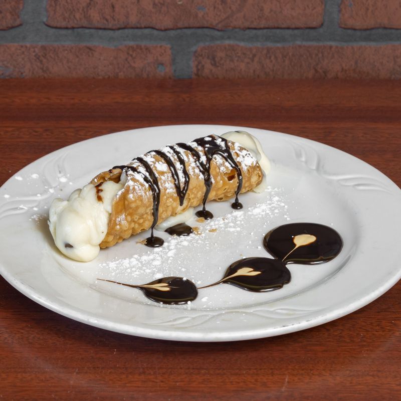 Cannoli on white plate, drizzled with chocolate, dusted with powdered sugar, on a wooden surface.
