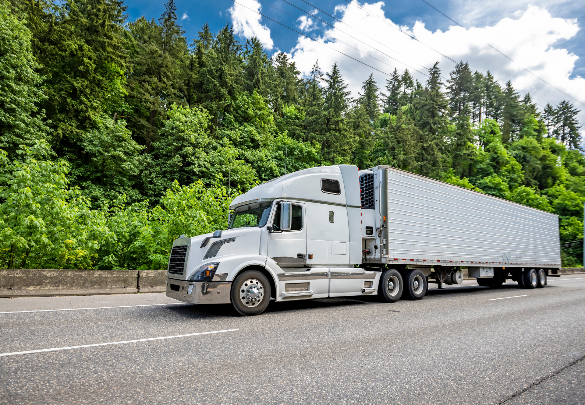 White semi-truck driving on a highway next to a lush green forest under a blue sky.