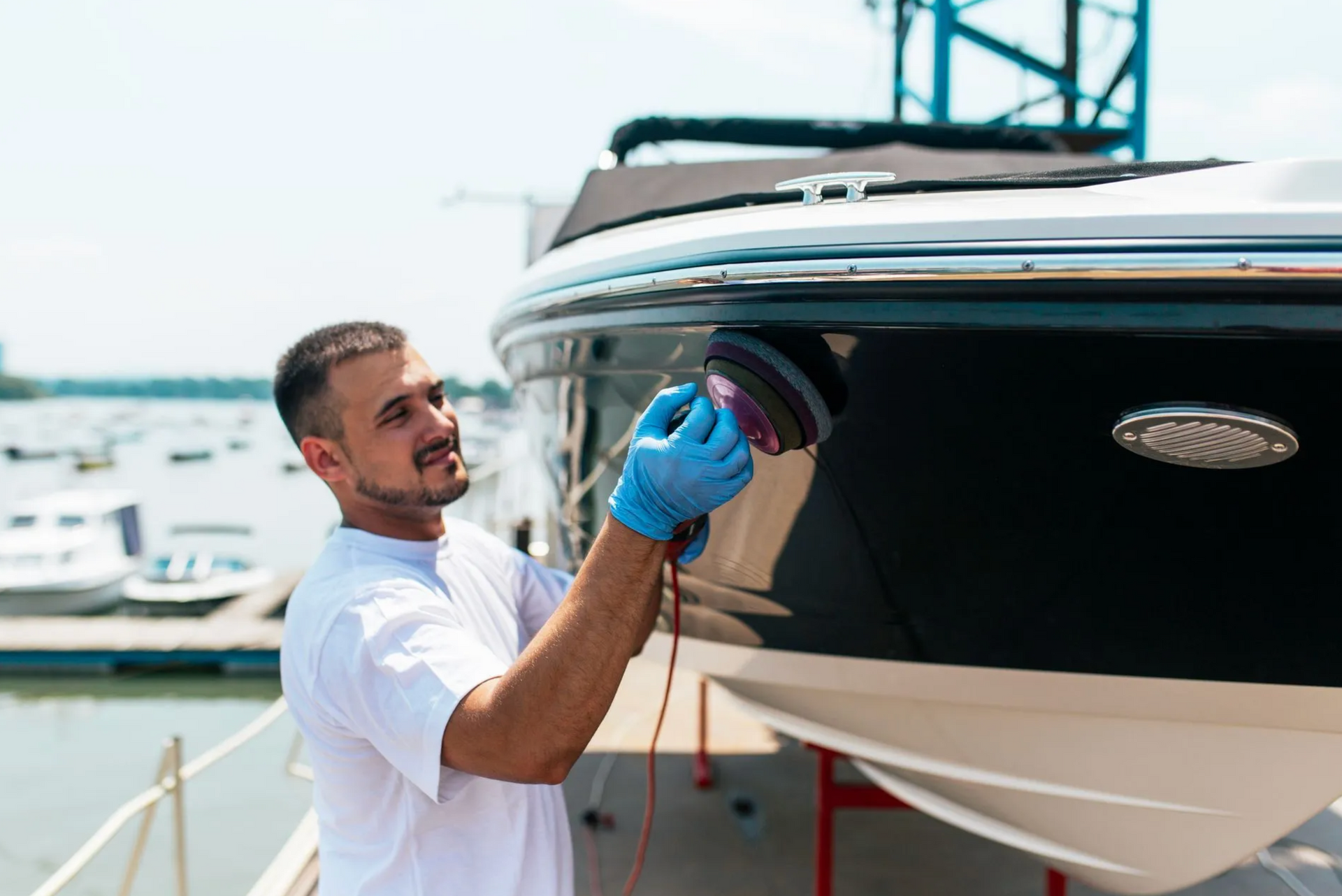 Man in blue gloves polishing a black and white boat, smiling in a sunny dockside setting.