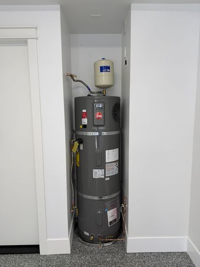 Water heater in a small closet alcove with exposed plumbing and a pressure tank above it