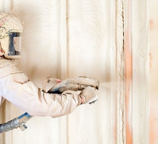 A man wearing a helmet and gloves is spraying foam on a wall.