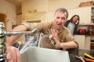 Man in flooded kitchen, water spraying from sink, holding a screwdriver; woman on phone in background.