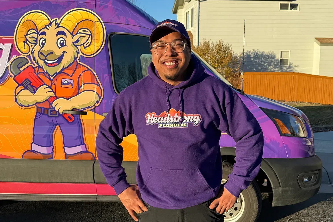 Man in purple hoodie stands in front of a van with a ram mascot holding a wrench.