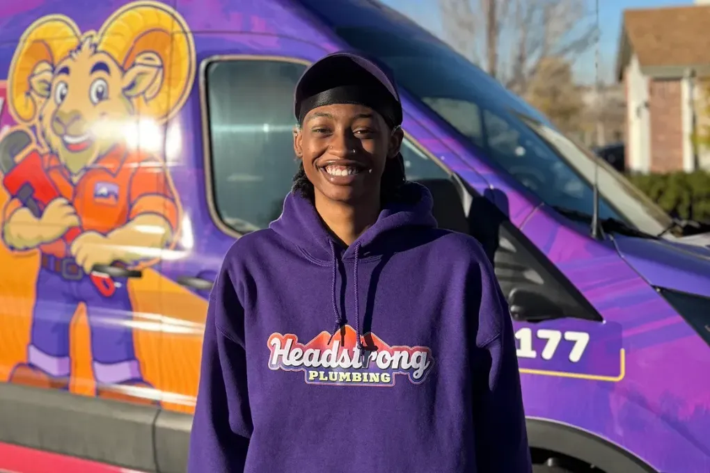 Person in purple hoodie smiling, standing in front of a purple van with a cartoon ram logo and the word 