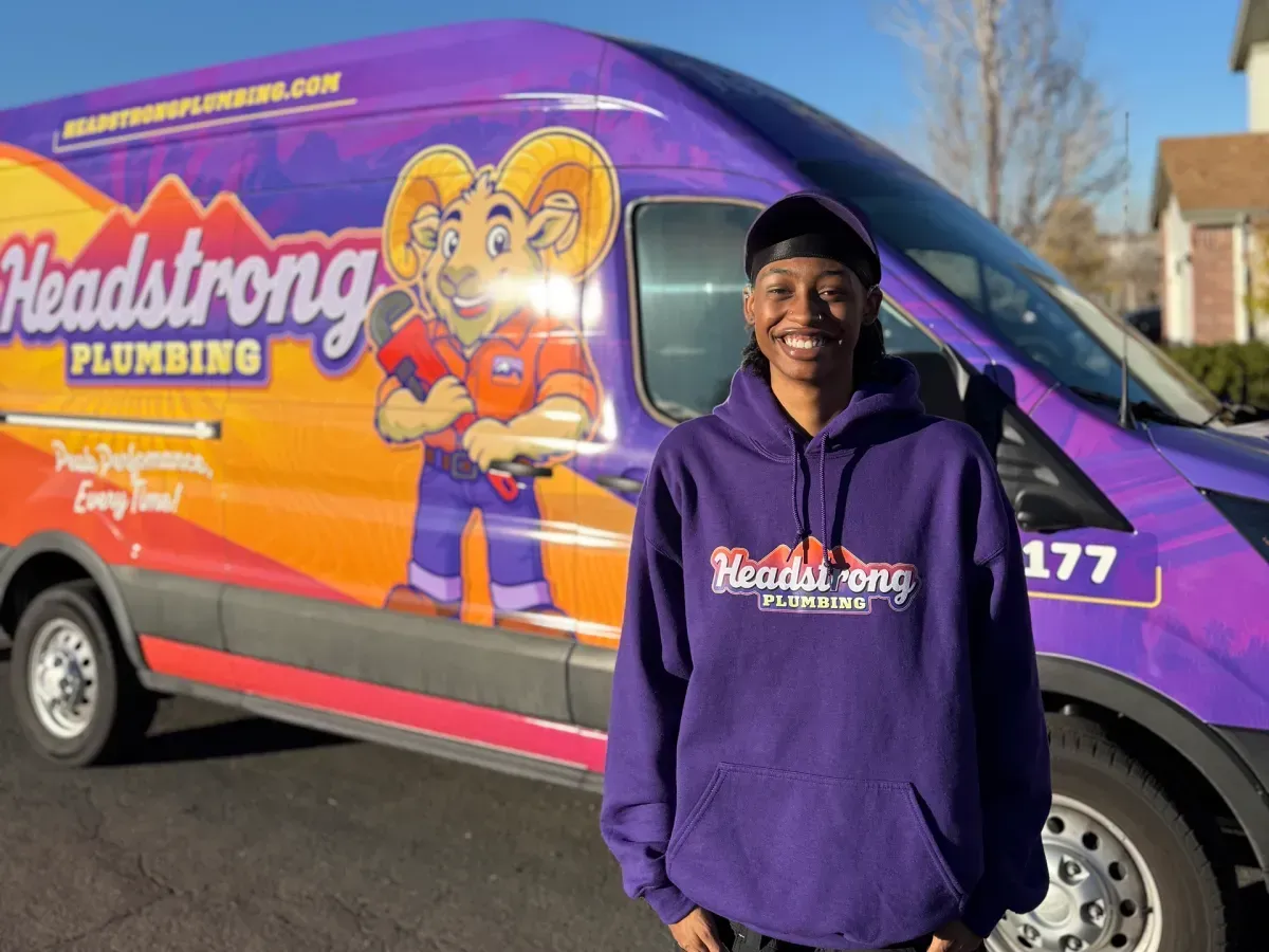 Person in purple hoodie stands near a Headstrong Plumbing van with a ram mascot in a residential setting.