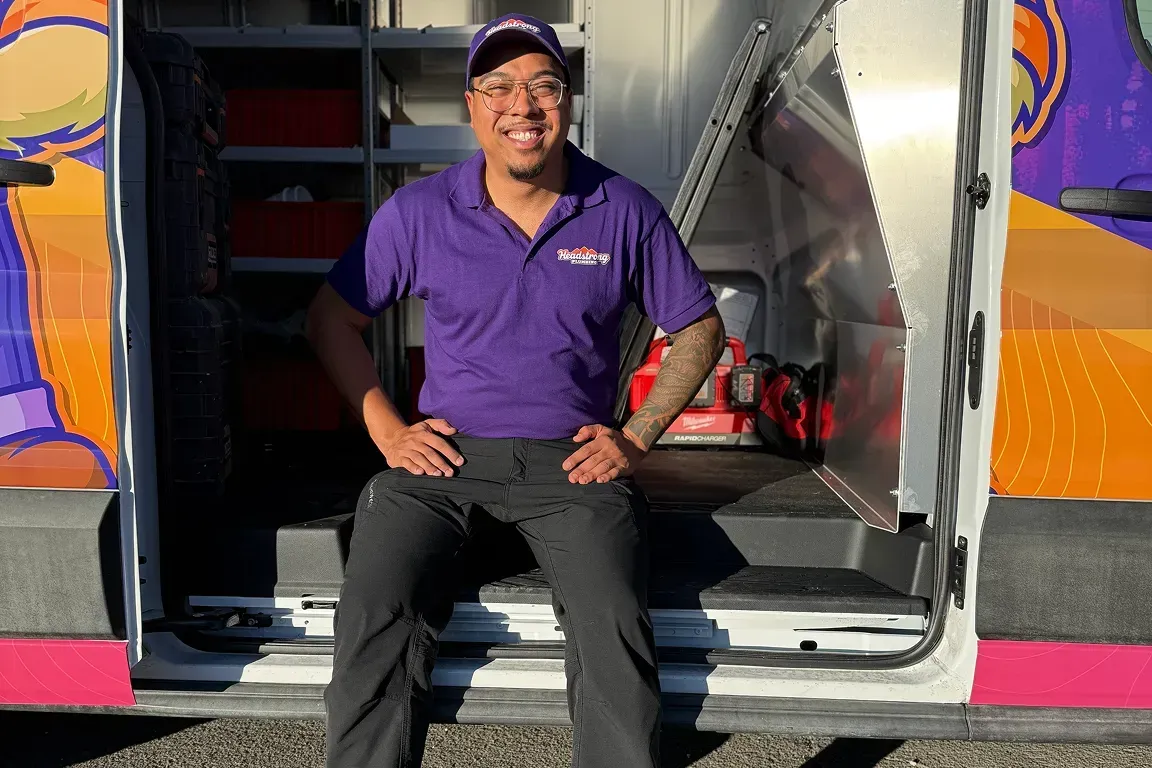 Smiling person in purple shirt sits in open van, hands on hips. Van has colorful mural.