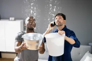 Two men catching water from a ceiling leak with buckets indoors. One is on the phone.