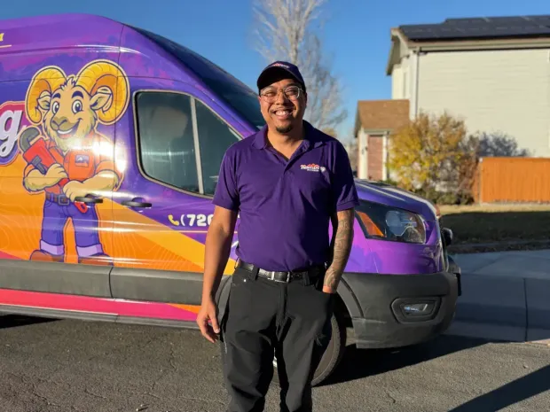 A smiling man in a purple shirt and hat stands in front of a purple van with a ram logo.