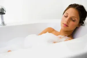 Woman relaxing in a bathtub filled with bubbles, eyes closed, white bathroom.