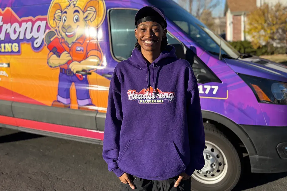 Person wearing purple hoodie stands in front of a van with a cartoon ram logo; sunny outdoor setting.
