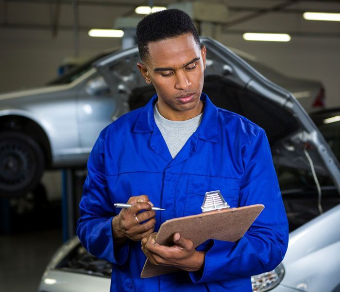 A mechanic is writing on a clipboard in front of a car with the hood open.