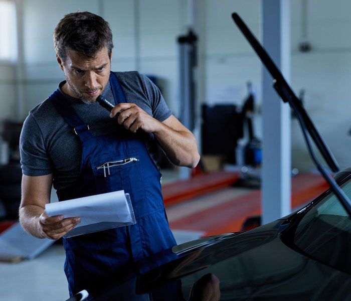 A man in blue overalls is looking at a piece of paper in front of a car.