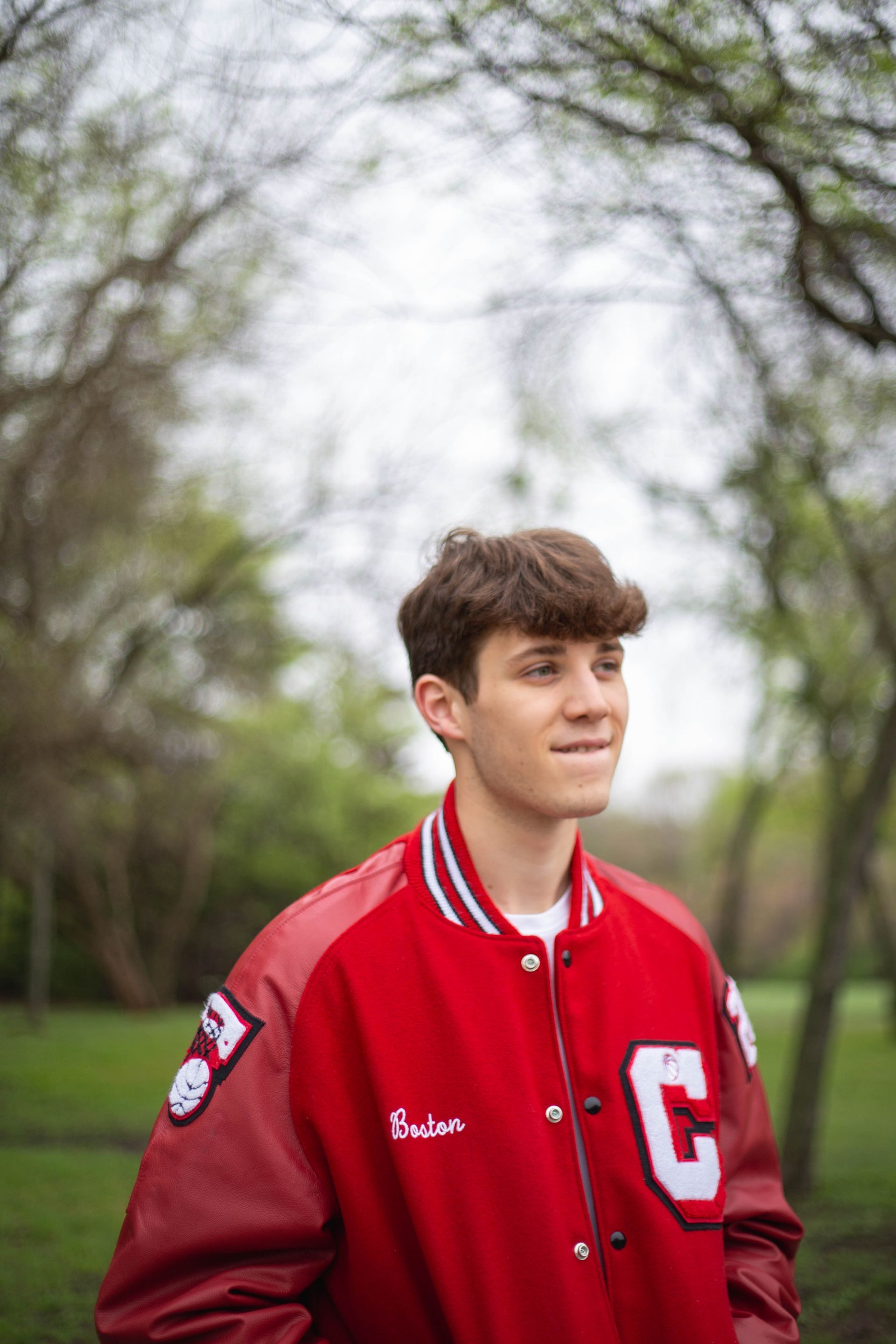 A young man wearing a red letterman jacket is standing in a park. Senior Photos by Sarah Rosie Studios.