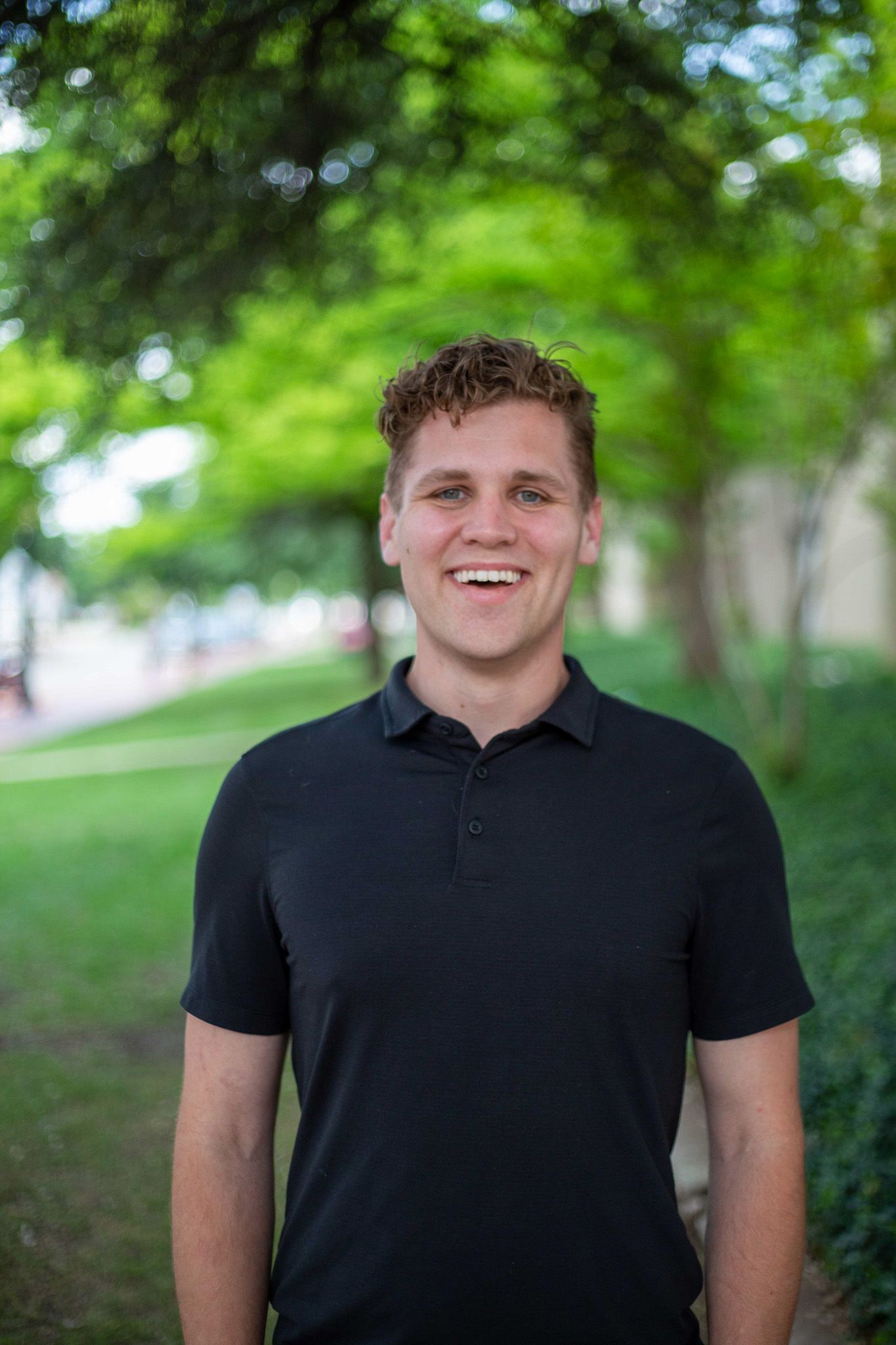 A young man in a black shirt is standing in a park. Corporate headshots by Sarah Rosie Studios.