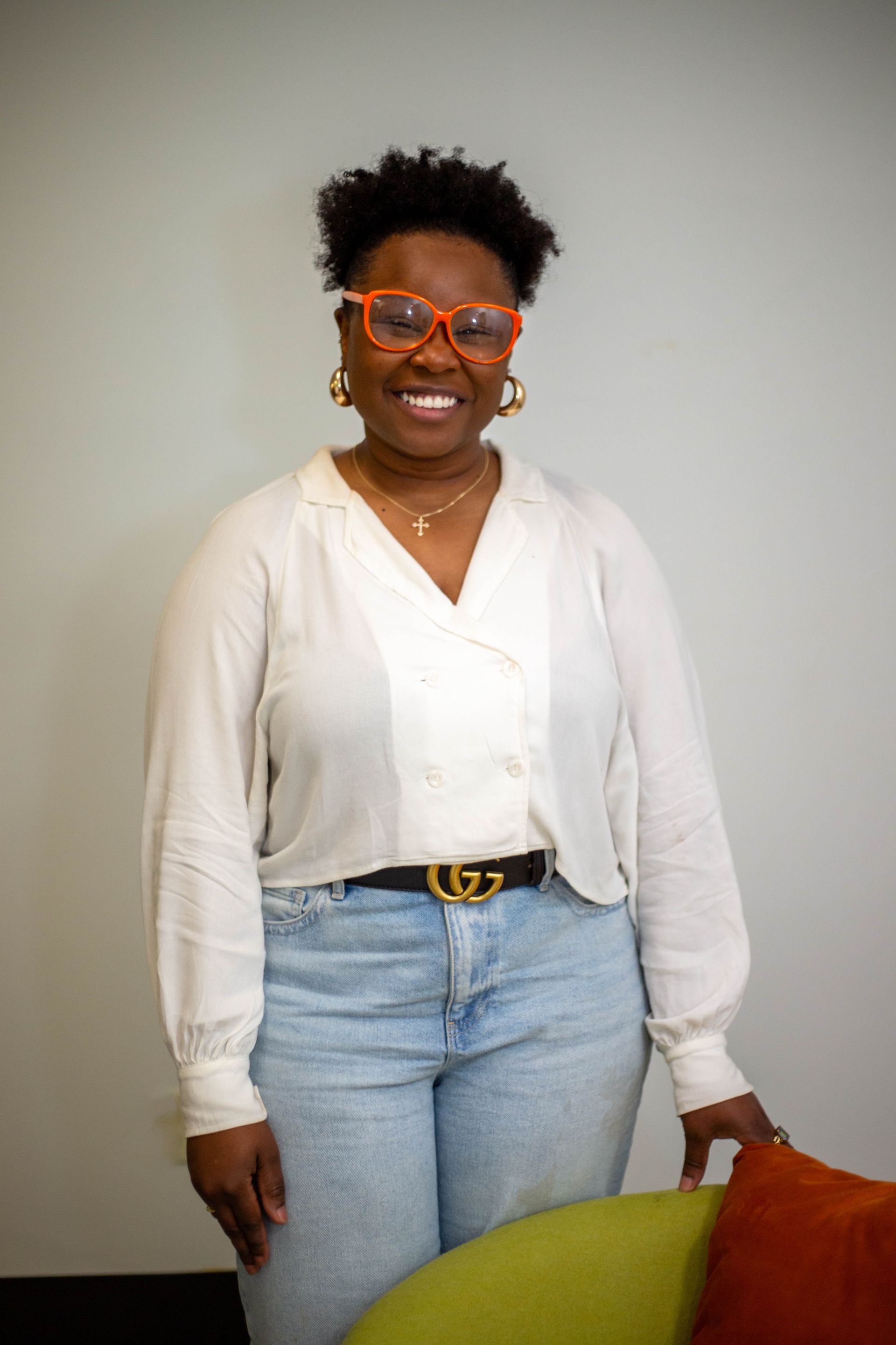 A woman wearing glasses and a white shirt is standing next to a green chair. Professional Corporate Headshots near you in Dallas, TX by Sarah Rosie Studios.