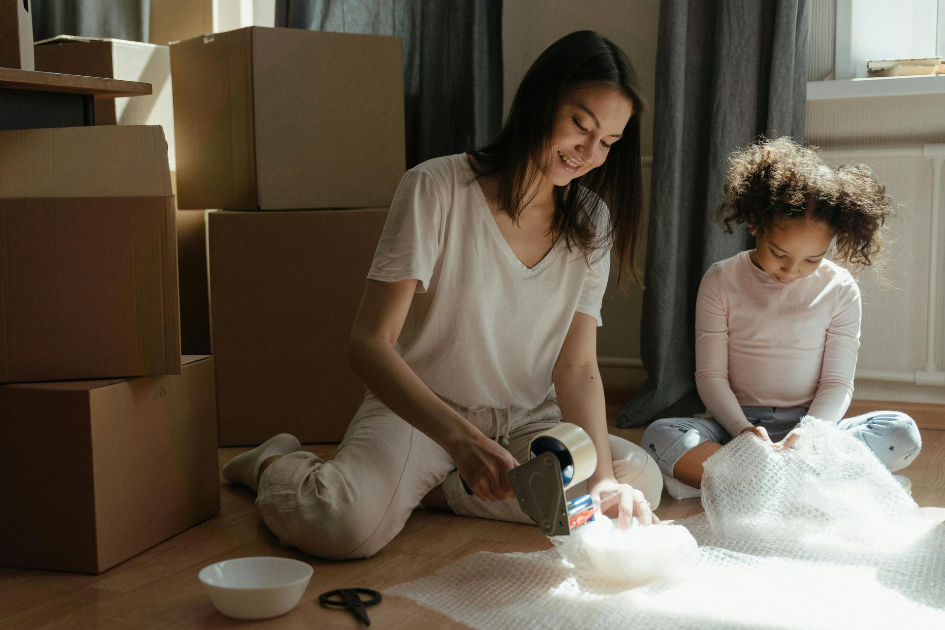 Image shows a single mother sitting on the floor packing a moving box with her daughter. 