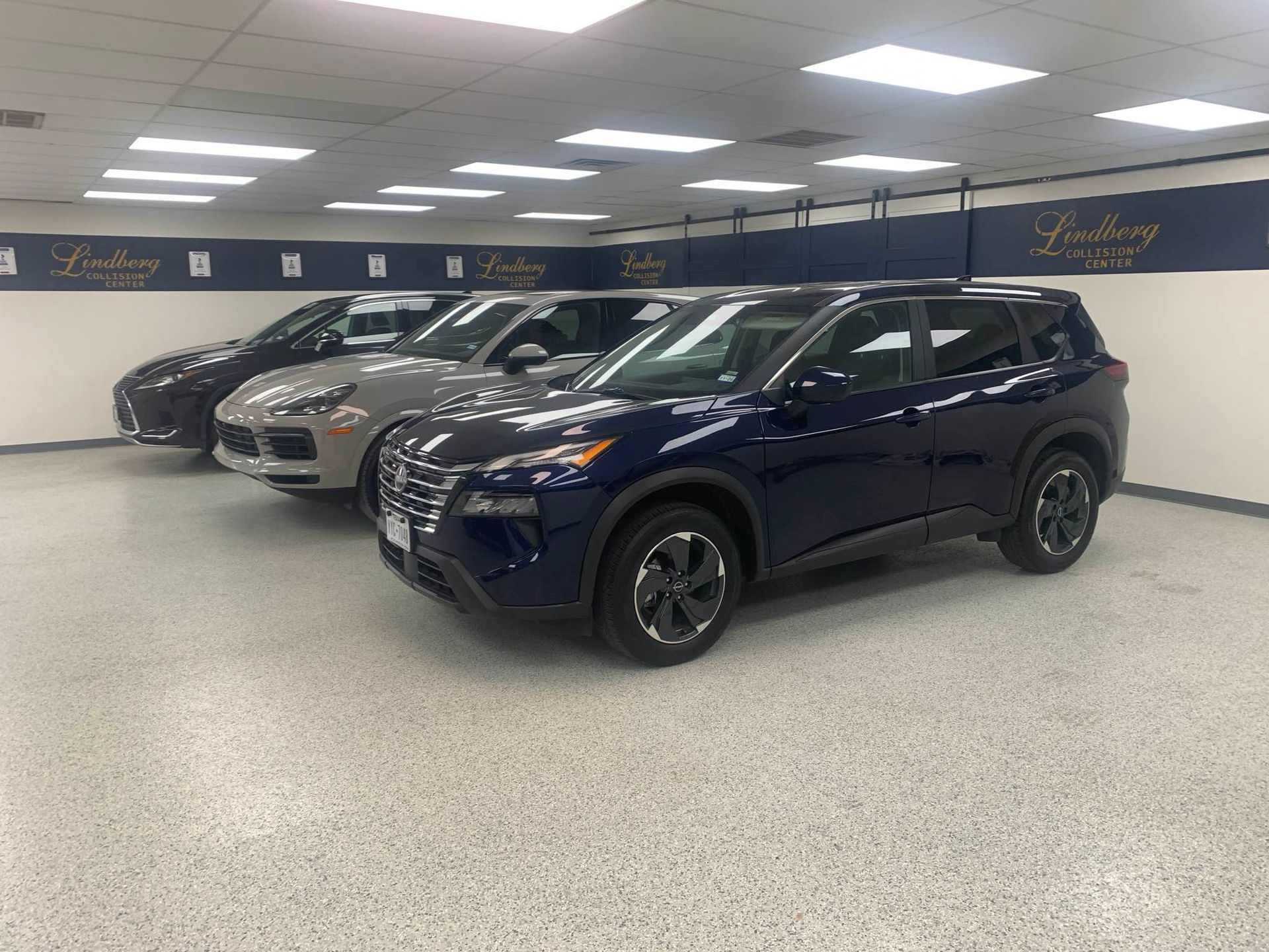 Row of SUVs in a dealership showroom: dark blue, gray, and black. Bright lights and logo on wall.
