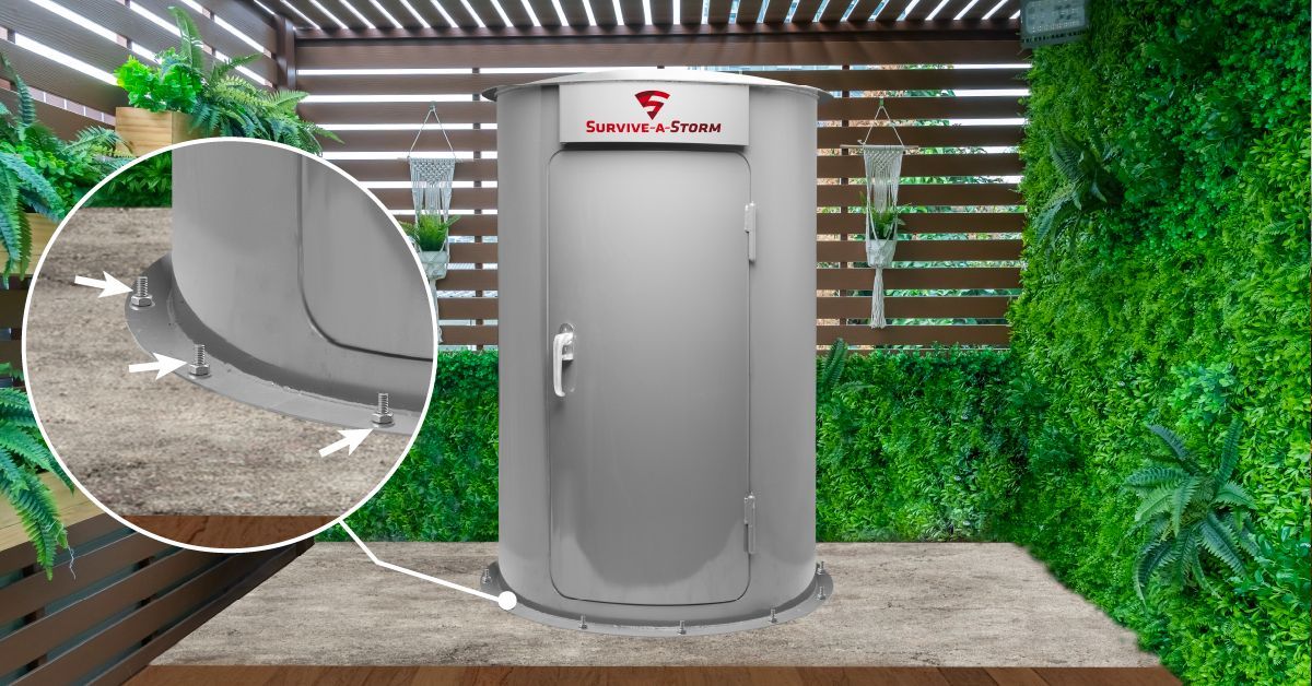 A grey, cylindrical outdoor tornado shelter stands on a stone patio with a callout highlighting its floor-anchoring bolts.