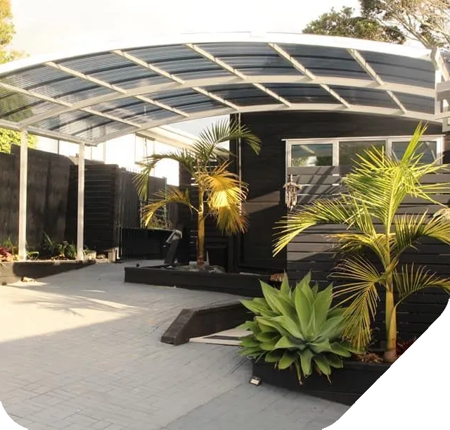 Carport with a transparent roof, attached to a black building, with surrounding greenery.