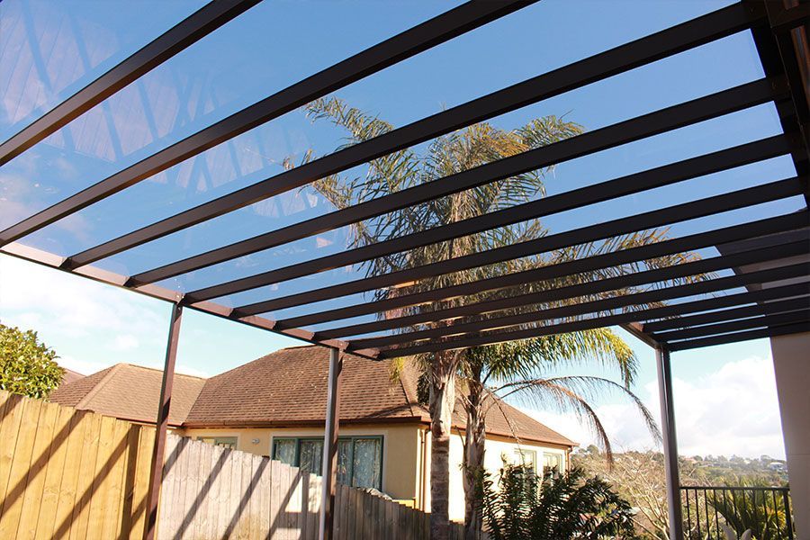 Pergola with translucent roof panels against a blue sky, over a house with a brown roof and wooden fence.