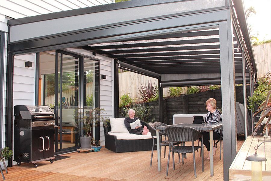 Two people relax under a black pergola on a wooden deck, beside a home with sliding glass doors.