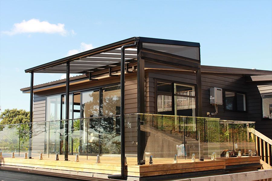 Wooden deck with glass railing, black pergola, and a house with glass windows on a sunny day.