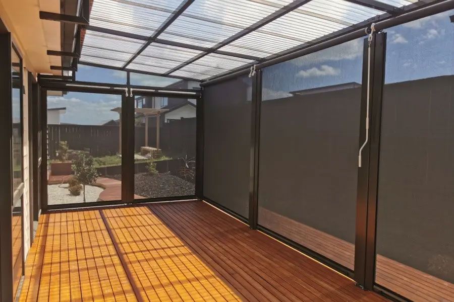 Sunroom with wooden deck, black framed glass windows and tinted shades. Translucent roof lets in sunlight.