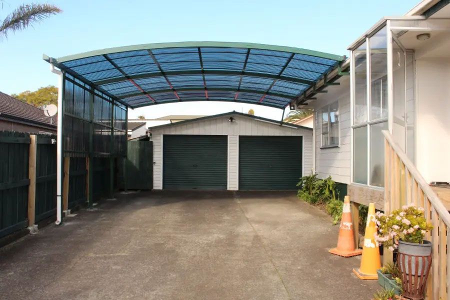 Carport with blue roof over driveway leading to a garage with two dark green doors.