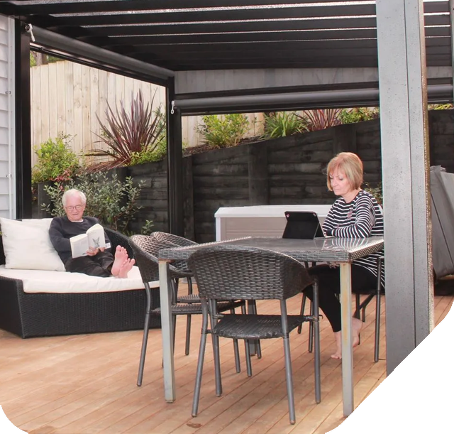 Two people relaxing under a pergola on a deck with a grill.