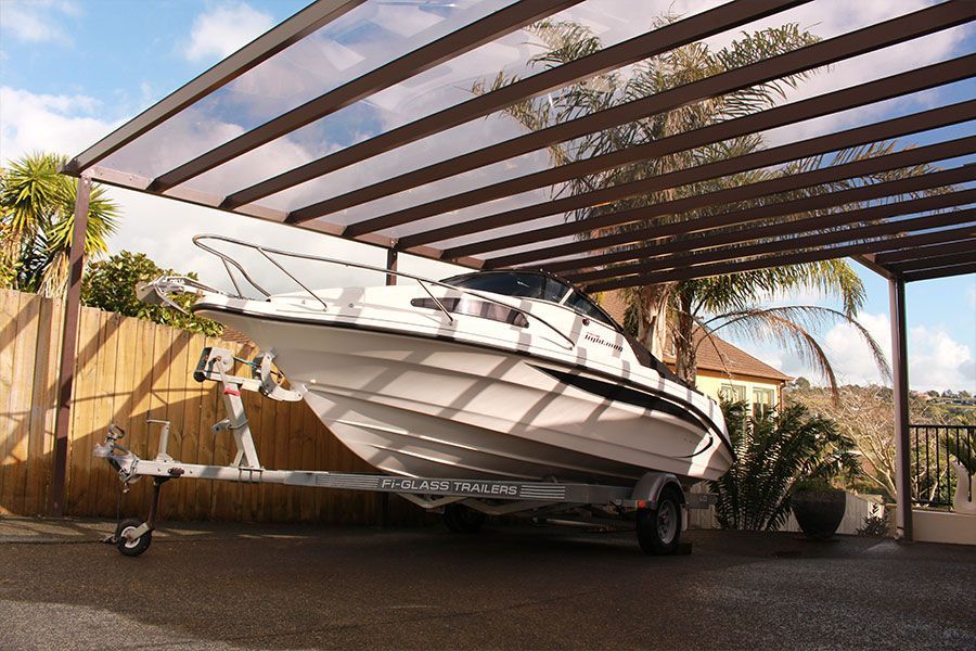 Boat on a trailer parked under a brown-framed shelter with a translucent roof, next to a bamboo fence.