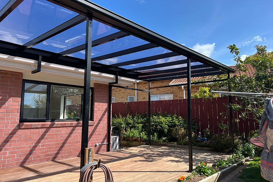 Black-framed pergola with clear roof over a brick patio. Bright blue sky background.